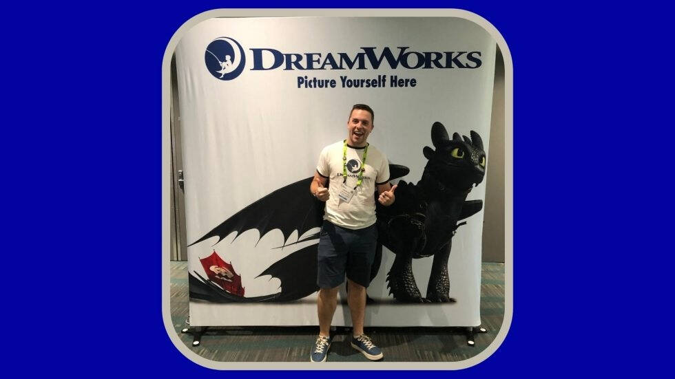 A person is standing in front of a DreamWorks promotional backdrop featuring Toothless, the dragon from "How to Train Your Dragon." The backdrop has the DreamWorks logo and the phrase "Picture Yourself Here." The person is giving two thumbs up.